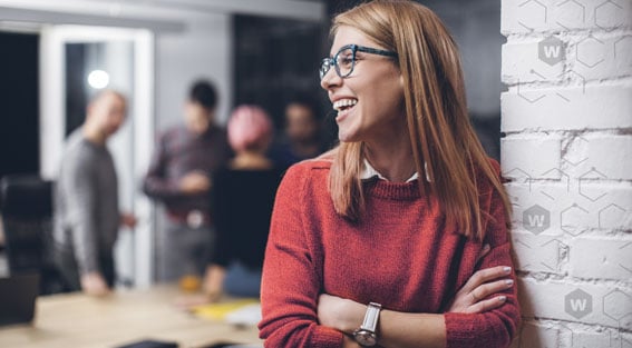 Smiling blonde in green glasses and a red sweater leaning against a white brick wall