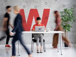 Woman working on her laptop at a white table with blurred people walking around her